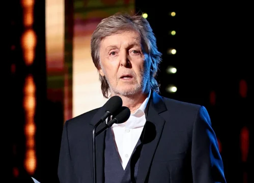 Older man in a dark suit speaks at a microphone on a stage with colorful lights.