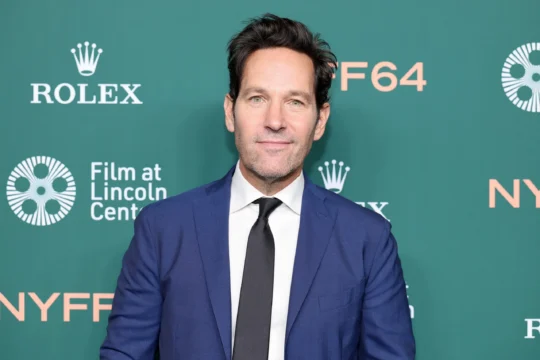 A man in a blue suit and black tie stands before a green backdrop with Rolex and NYFF logos.