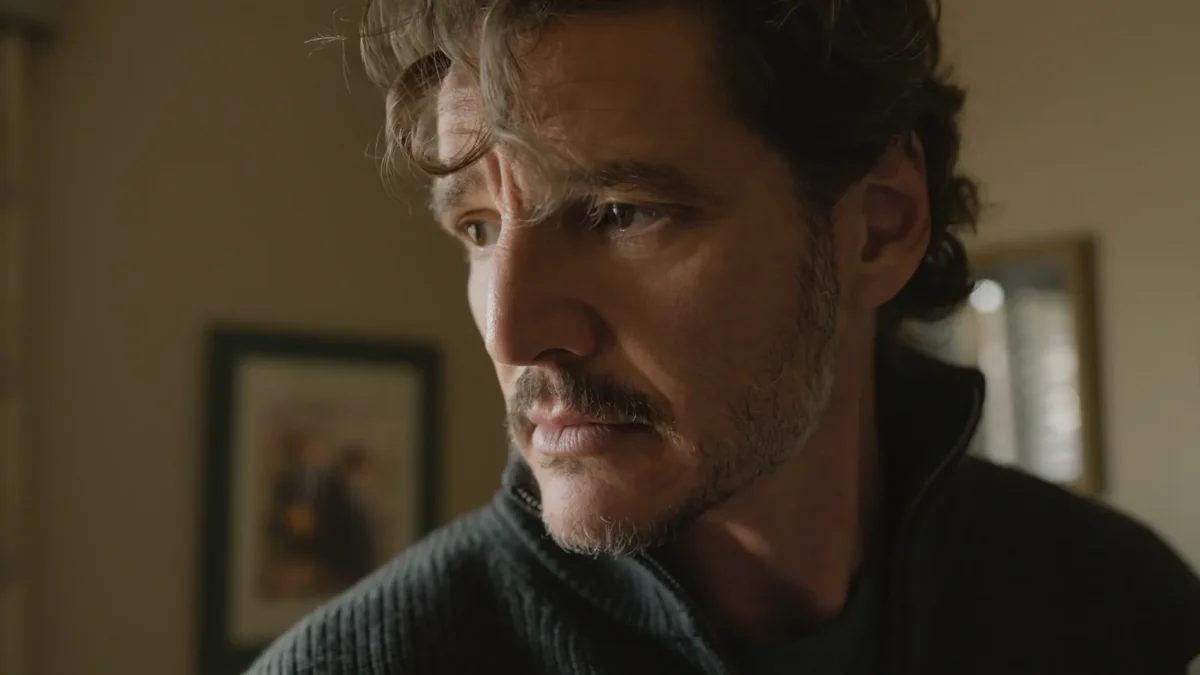 A man with wavy hair and stubble looks intently to the side in a dimly lit room.