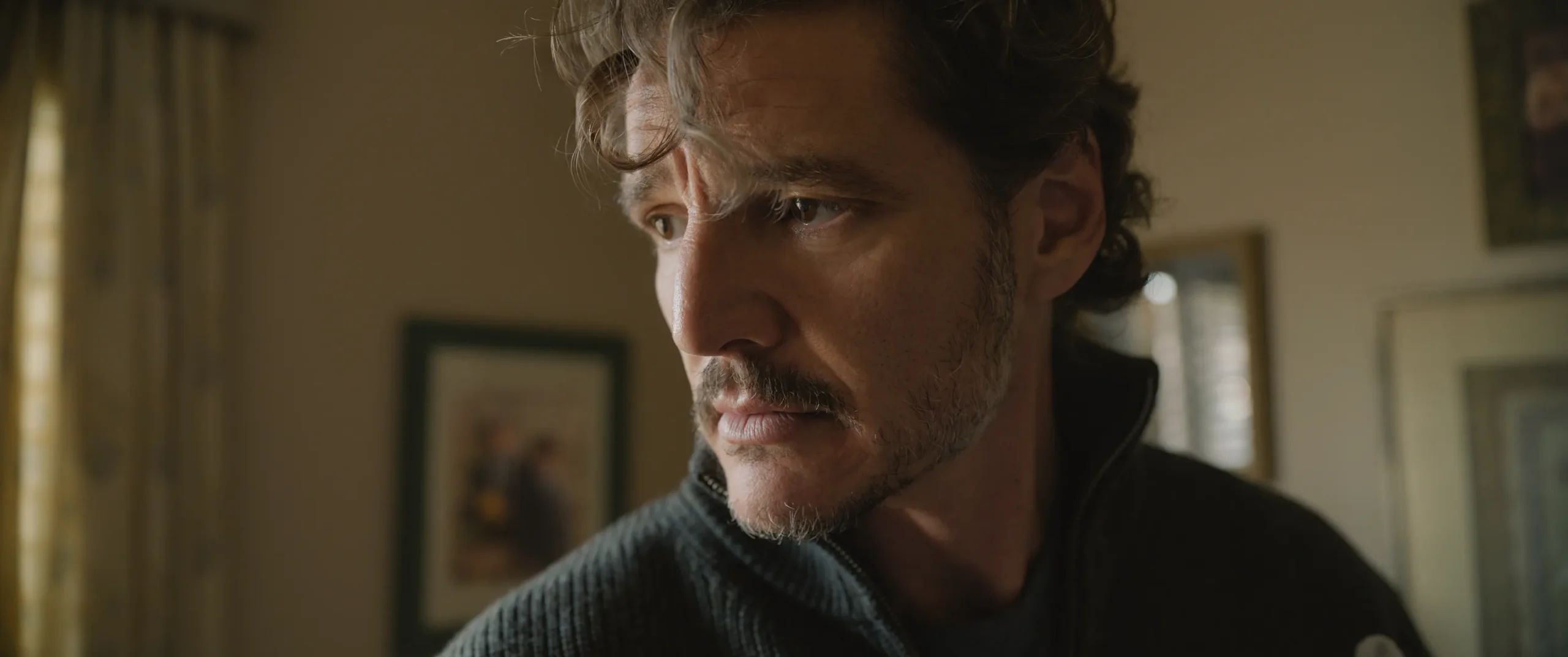 A man with wavy hair and stubble looks intently to the side in a dimly lit room.