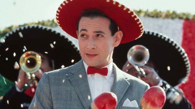A man in a gray suit and red sombrero stands before musicians playing brass instruments.