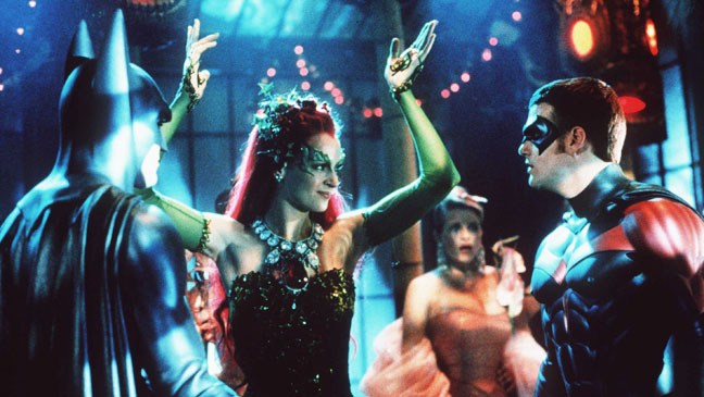 A green-costumed woman raises her arms between two masked, caped figures in a colorful party scene.