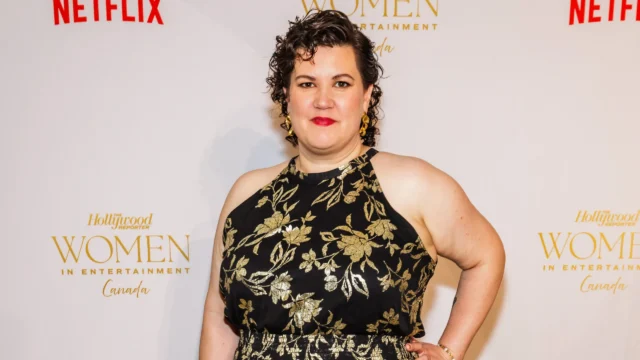 A woman in a black dress with gold floral patterns poses before a logo-covered backdrop.