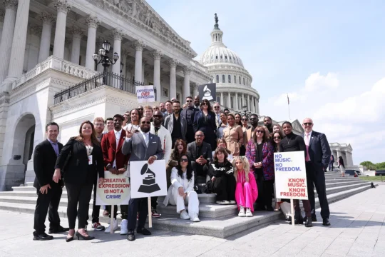 A large group poses on steps outside a domed neoclassical building, holding signs about music and expression.