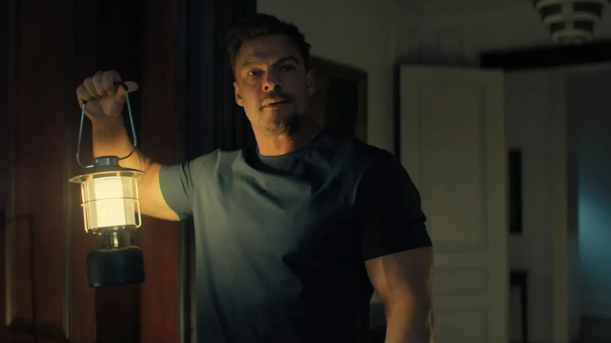A man in a dark T-shirt holds a glowing lantern in a dimly lit room.