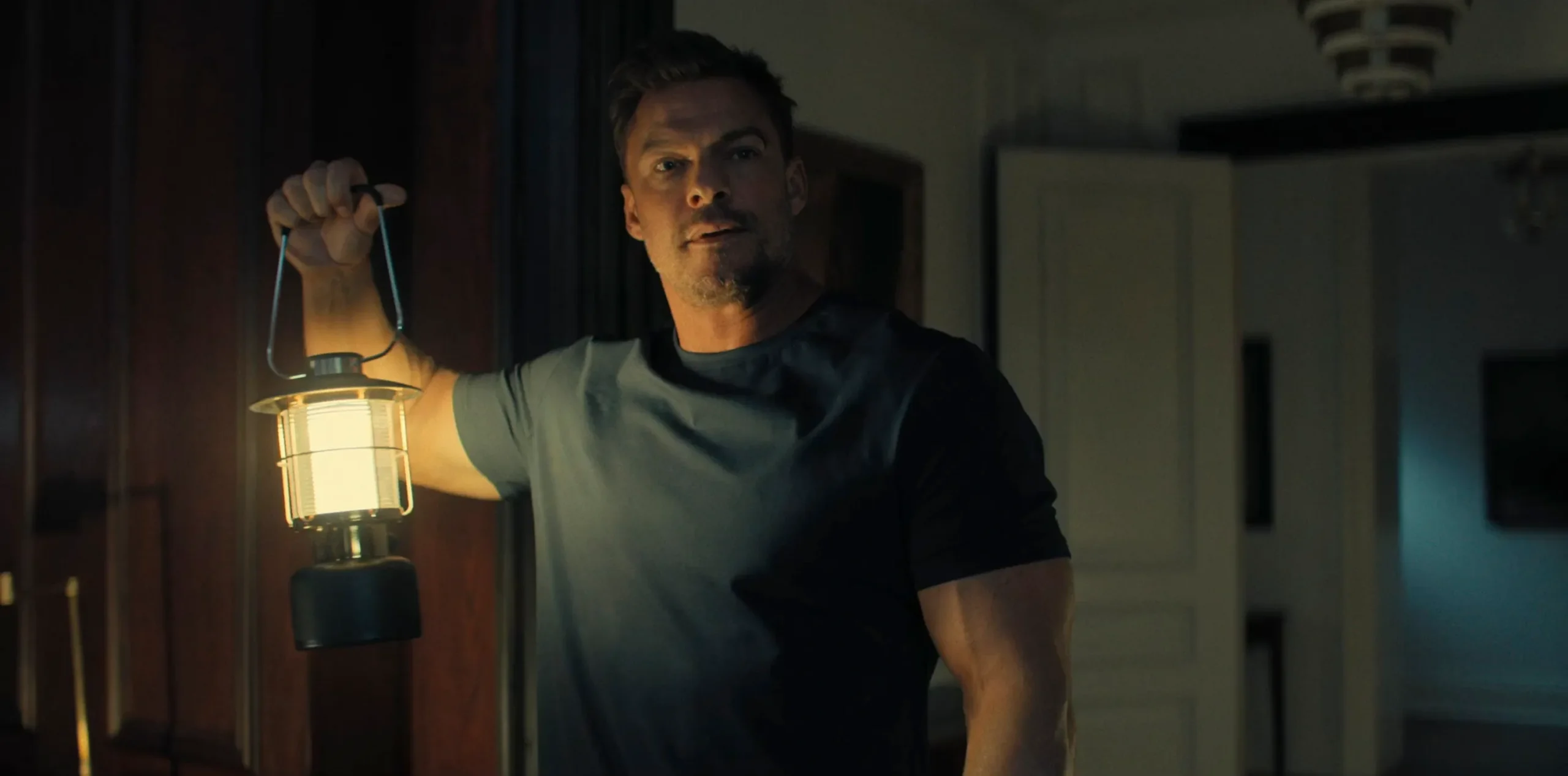 A man in a dark T-shirt holds a glowing lantern in a dimly lit room.