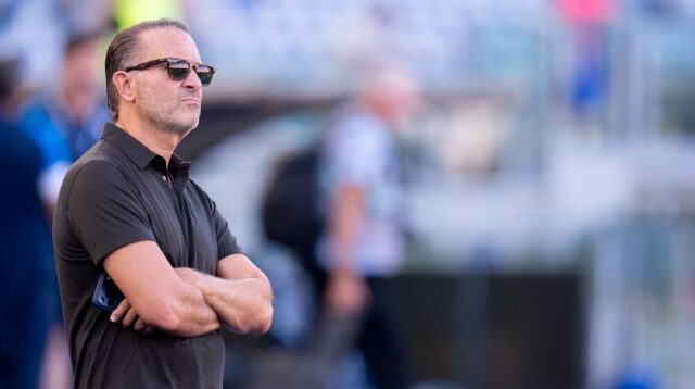 A man in sunglasses and a black polo stands with arms crossed against a blurred background.