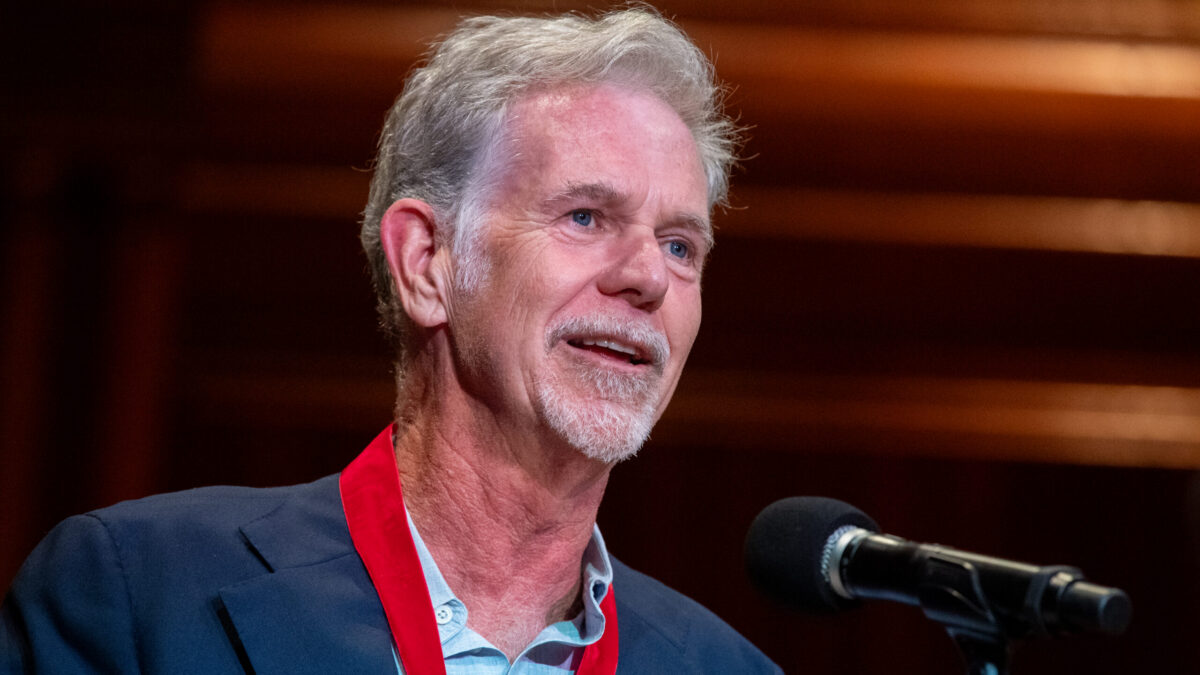 Gray-haired man in a blue blazer and red lanyard sits beside a microphone against a wood-paneled background.