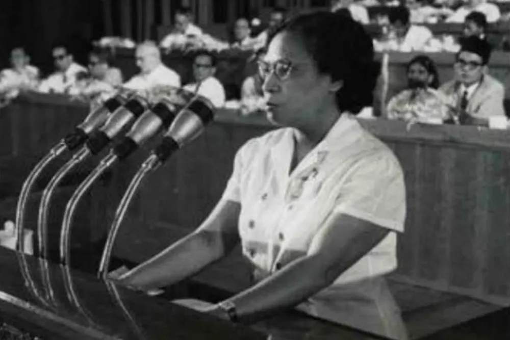 A woman in glasses stands at a podium with several microphones before seated attendees.