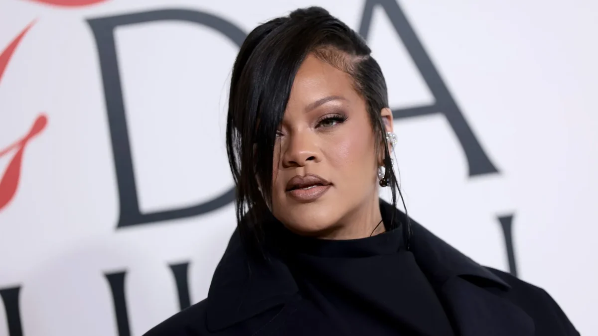 Woman with side-swept dark hair and earrings poses in a black outfit before a large-lettered backdrop.