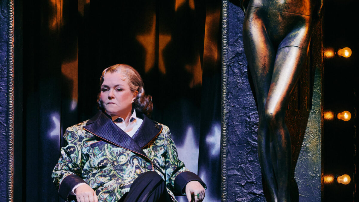 A stern-looking performer sits in an ornate chair beside a bronze nude statue and glowing stage lights.