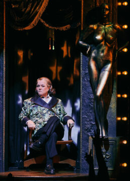 A stern-looking performer sits in an ornate chair beside a bronze nude statue and glowing stage lights.