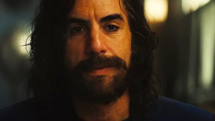 A bearded man with long dark hair looks forward in dim, warm lighting.