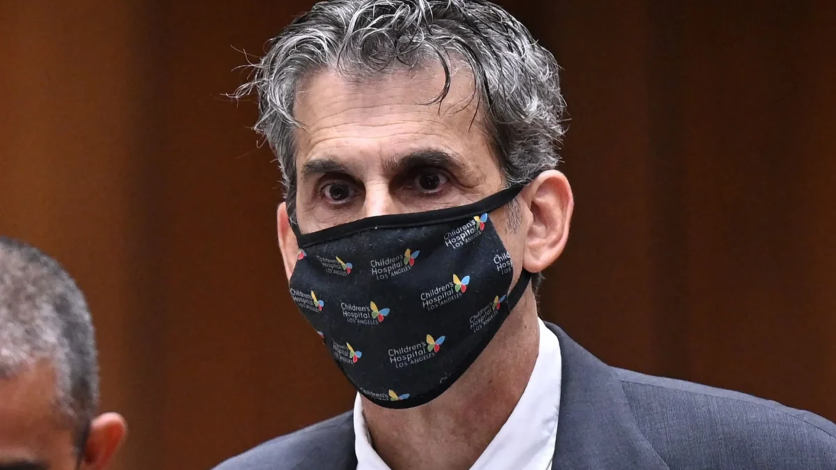 A gray-haired man in a suit wears a black Children's Hospital Los Angeles face mask against a wood-paneled background.
