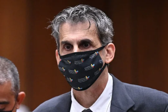 A gray-haired man in a suit wears a black Children's Hospital Los Angeles face mask against a wood-paneled background.
