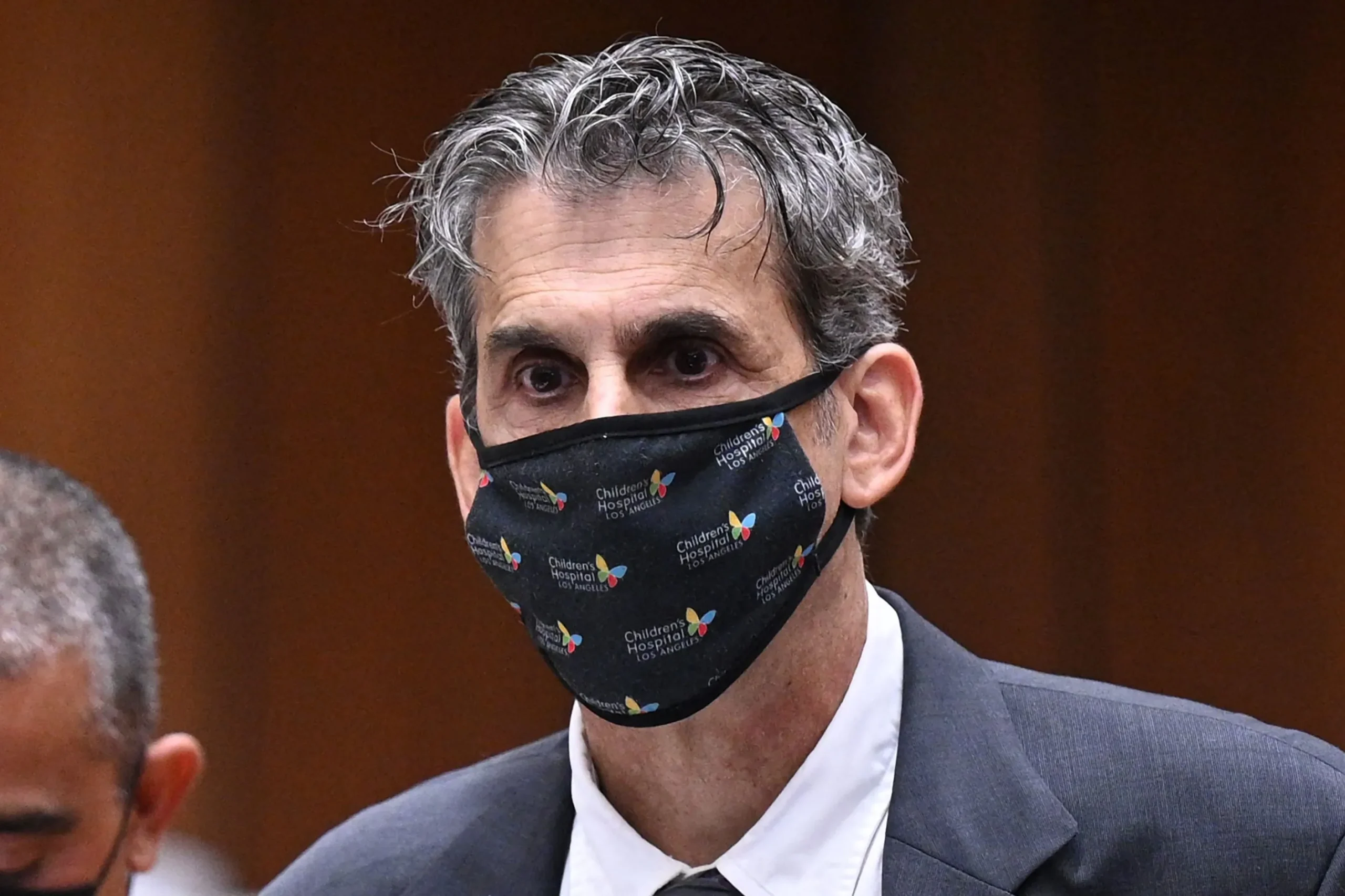 A gray-haired man in a suit wears a black Children's Hospital Los Angeles face mask against a wood-paneled background.