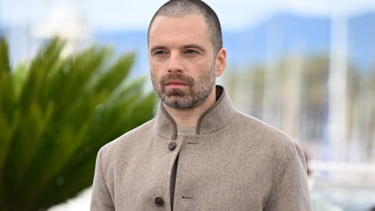 A man with a buzz cut and beard wears a beige buttoned coat outdoors against a blurred background.