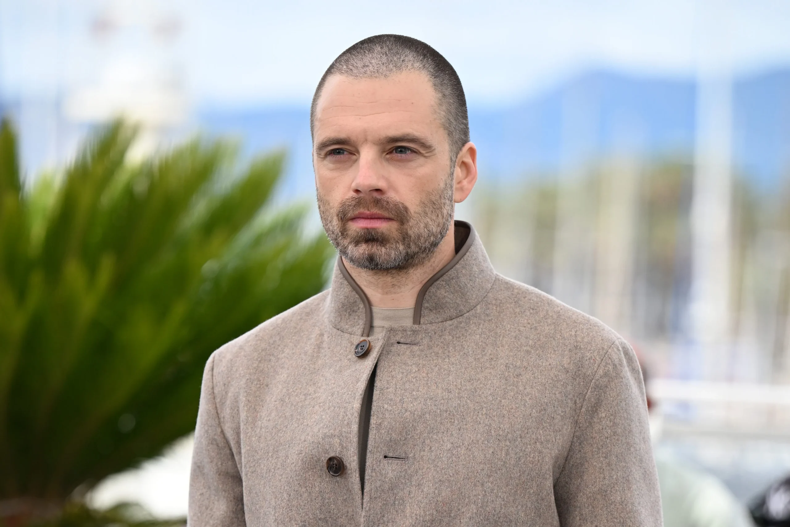 A man with a buzz cut and beard wears a beige buttoned coat outdoors against a blurred background.