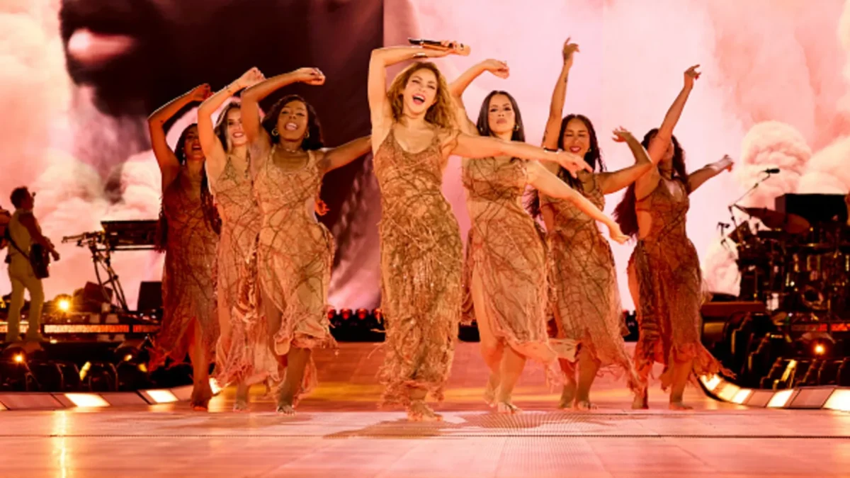 A performer holding a microphone dances with several performers in gold dresses on a brightly lit stage.