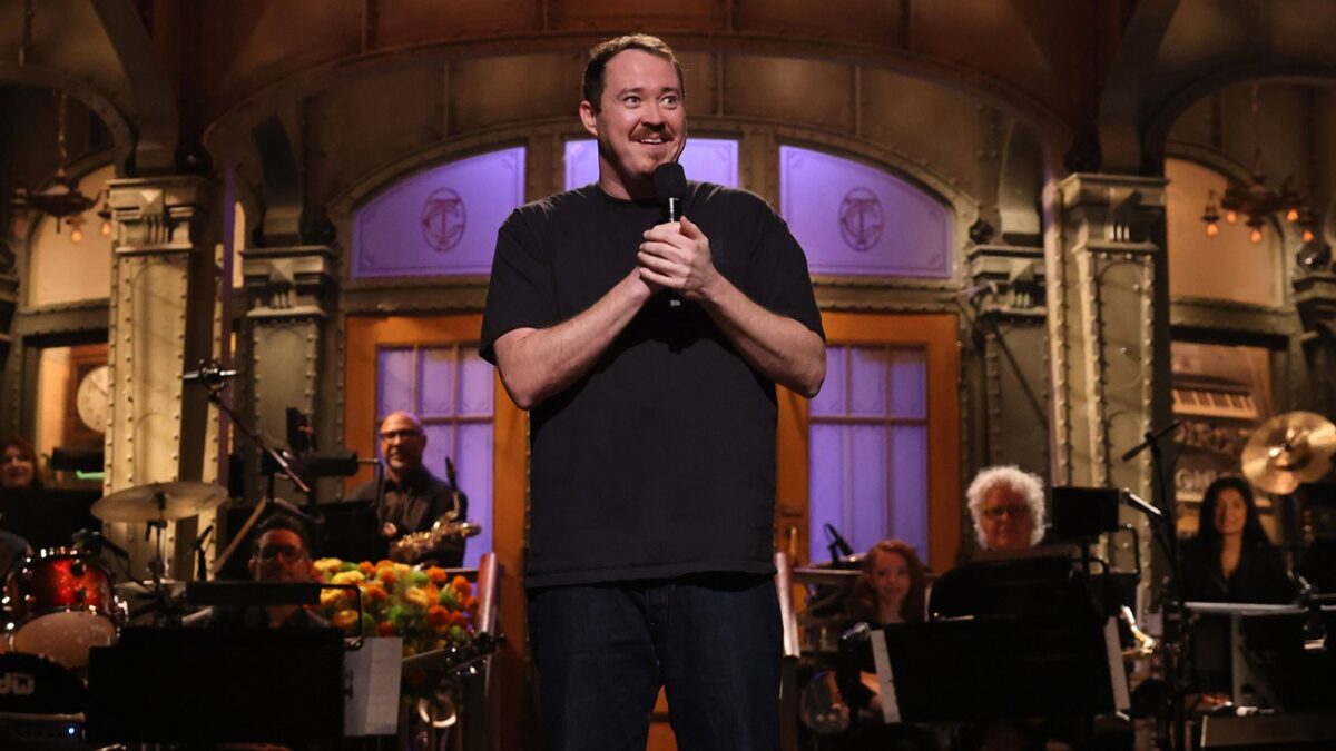 A man in a black shirt holds a microphone on a stage with musicians behind him.