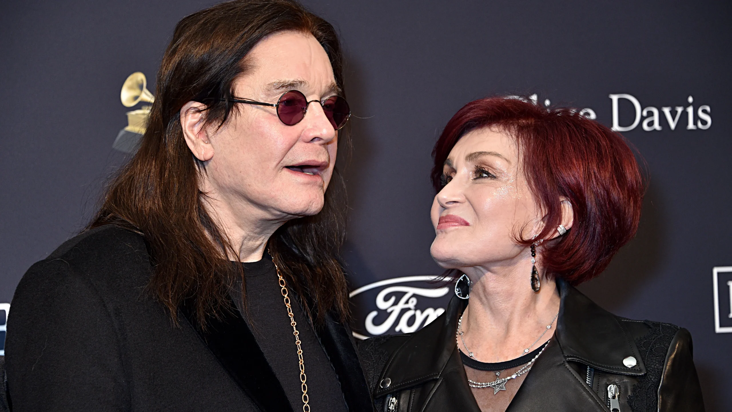 A long-haired man in round sunglasses stands beside a red-haired woman looking up at him.
