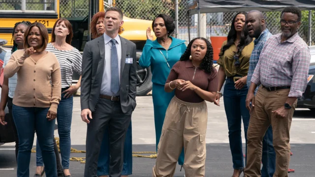 A group of adults stand outdoors near a yellow school bus, looking concerned toward something off-camera.