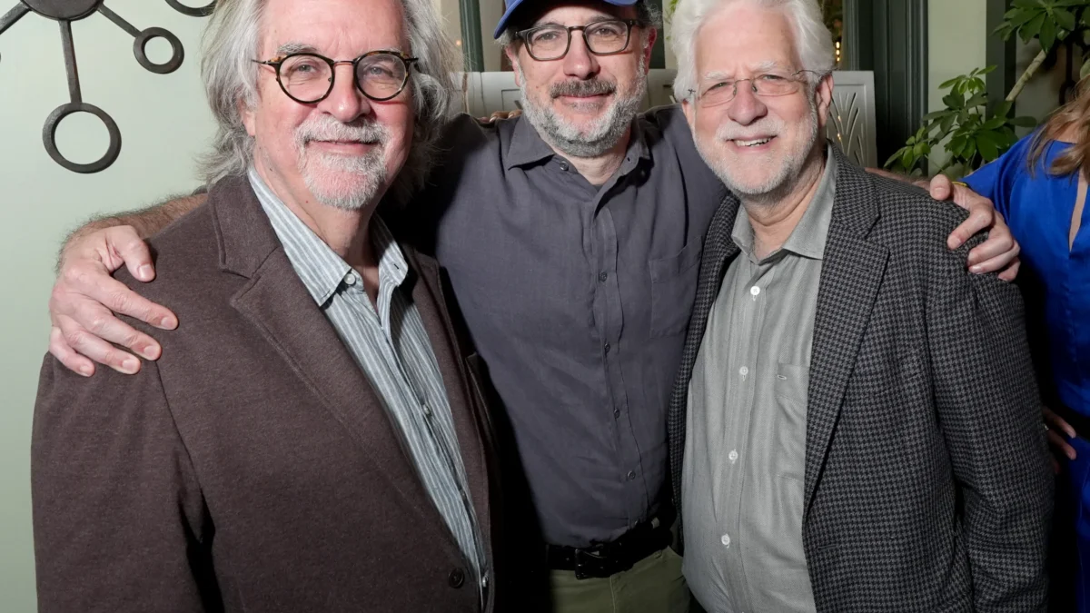 Three smiling older men with glasses pose with arms around each other indoors.