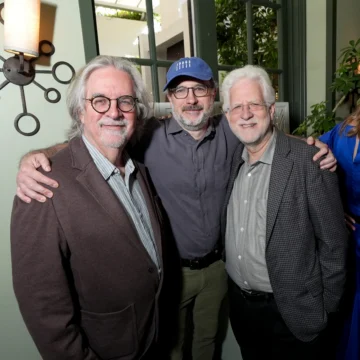 Three smiling older men with glasses pose with arms around each other indoors.