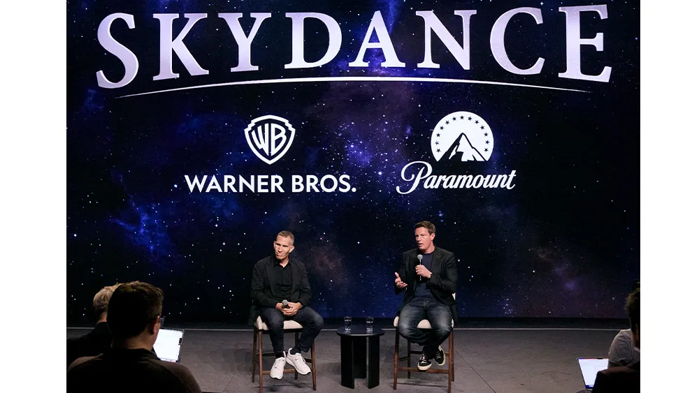 Two men sit with microphones onstage before a starry screen displaying Skydance, Warner Bros., and Paramount logos.