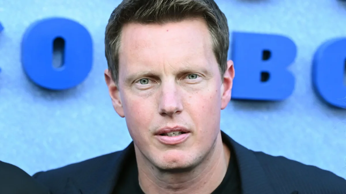 Close-up of a man in a dark jacket before large blue letters on a gray background.