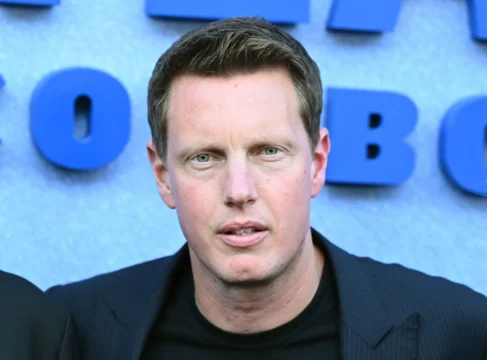 Close-up of a man in a dark jacket before large blue letters on a gray background.