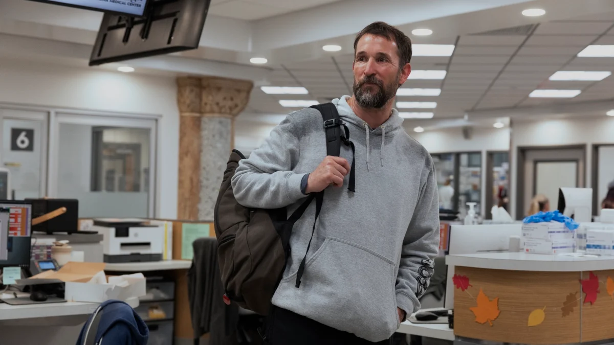 Bearded man in a gray hoodie carries a backpack through a hospital reception area.