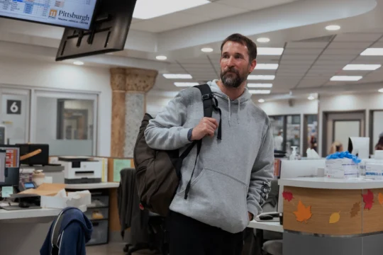 Bearded man in a gray hoodie carries a backpack through a hospital reception area.