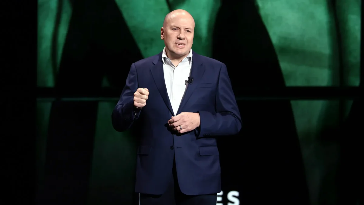 A bald man in a navy suit gestures while speaking on a dark stage with green lighting.