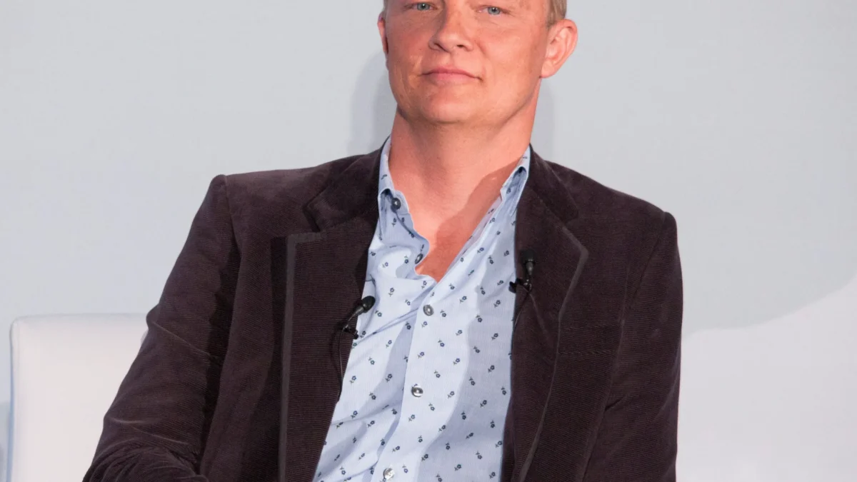 A man in a blazer sits in a white chair with microphones in front of a plain backdrop.