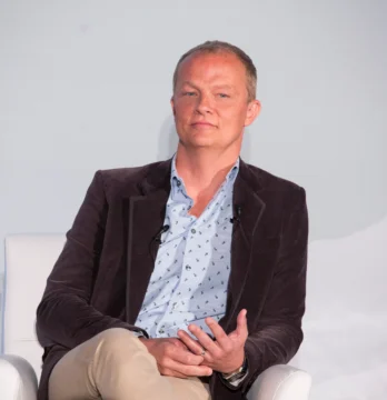 A man in a blazer sits in a white chair with microphones in front of a plain backdrop.