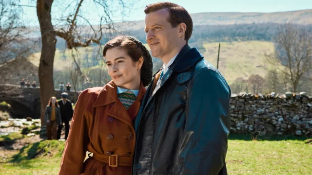 A woman in a brown coat and a man in a dark jacket stand together in a sunny rural landscape.