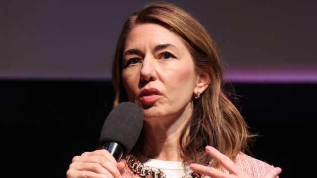 A woman with long brown hair speaks into a handheld microphone against a dark background.