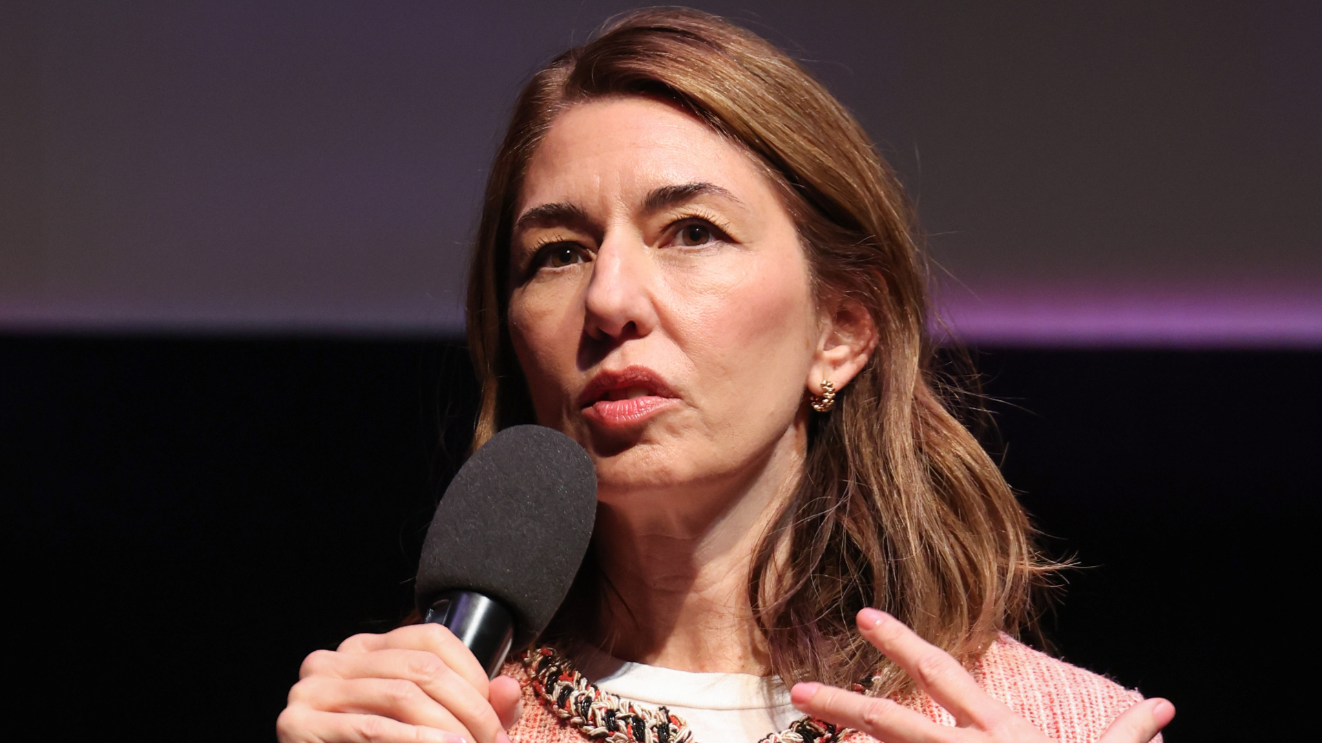 A woman with long brown hair speaks into a handheld microphone against a dark background.