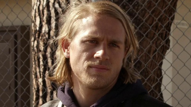Blond man with shoulder-length hair stands near a chain-link fence, looking off to the side.