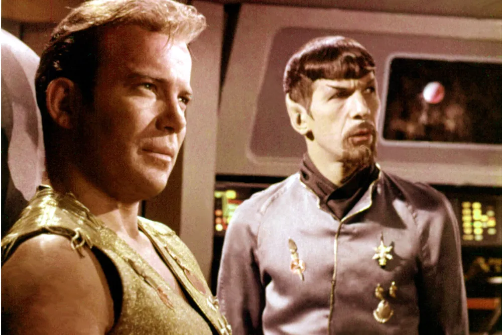 Two men in futuristic costumes stand on a spaceship-like set, one wearing pointed-ear makeup.