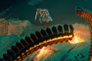 A large segmented appendage arcs over flames in a rocky scene, with a small armored figure in the background.