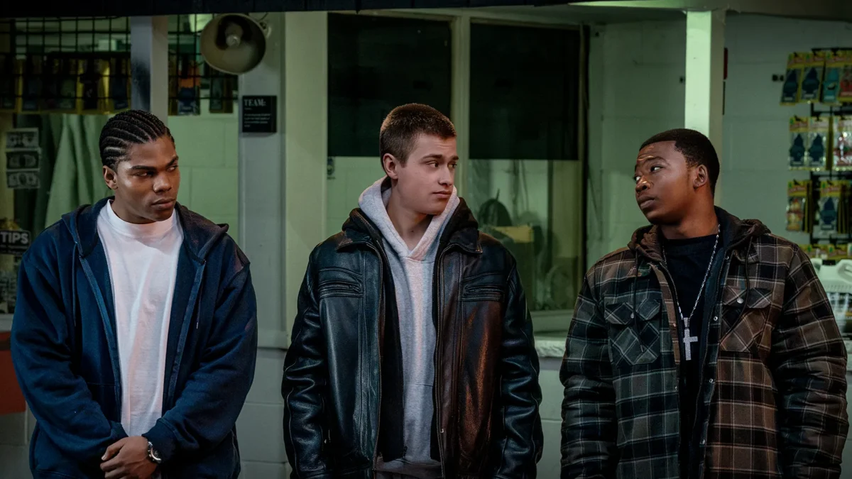Three young men in jackets stand indoors under dim greenish lighting, looking at one another.