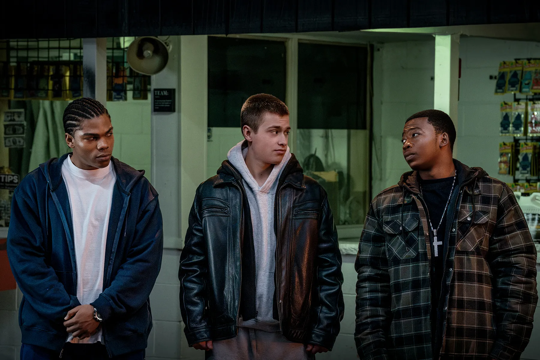 Three young men in jackets stand indoors under dim greenish lighting, looking at one another.