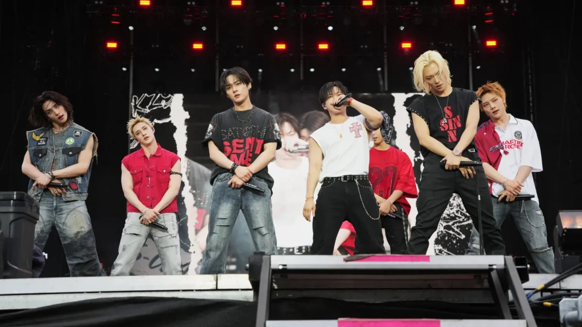 Seven performers holding microphones stand on a stage with red lights and a large screen behind them.