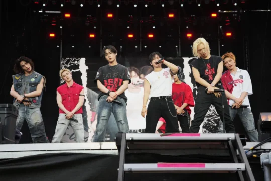 Seven performers holding microphones stand on a stage with red lights and a large screen behind them.