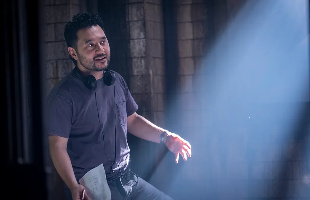A man with headphones around his neck holds papers beside a stone wall in dramatic blue light.