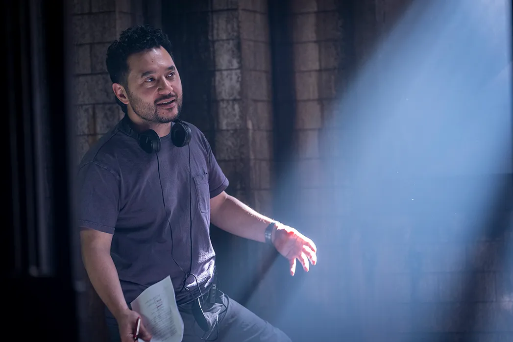 A man with headphones around his neck holds papers beside a stone wall in dramatic blue light.