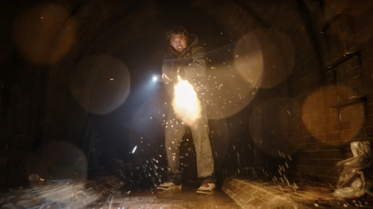 A person fires a flame-emitting device in a dark, wet brick corridor filled with sparks.