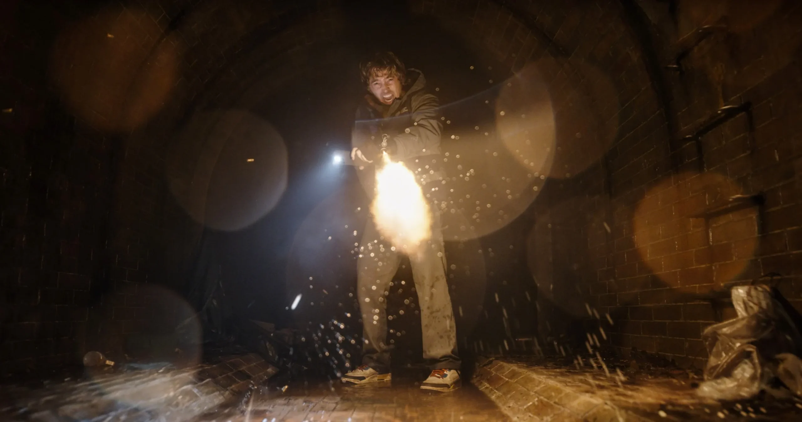 A person fires a flame-emitting device in a dark, wet brick corridor filled with sparks.
